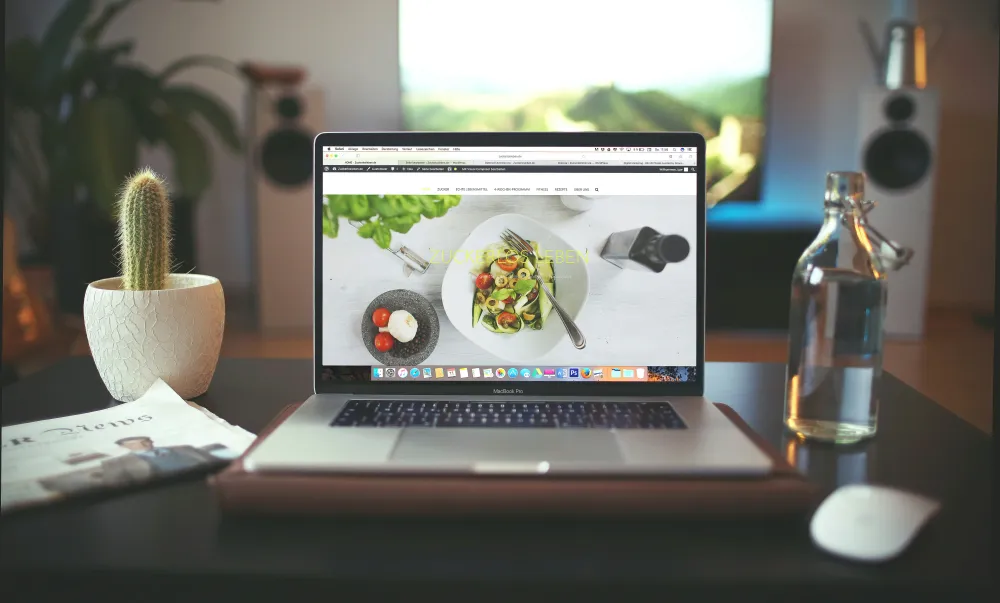 A laptop displays a salad recipe website, surrounded by a cactus, a glass bottle of water, and a magazine on a dark table.