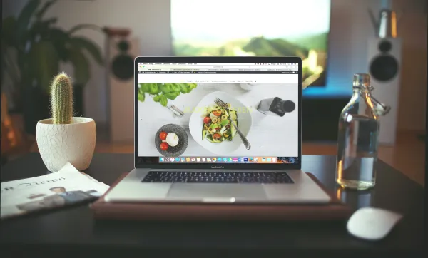 A laptop displays a salad recipe website, surrounded by a cactus, a glass bottle of water, and a magazine on a dark table.